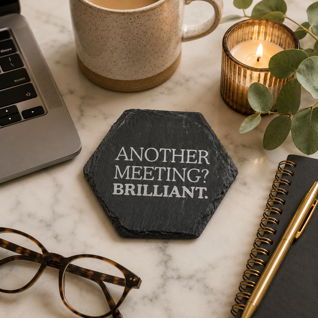Hexagonal slate coaster with 'Another meeting? Brilliant.' text on a marble desk with laptop, notebook, and pens.