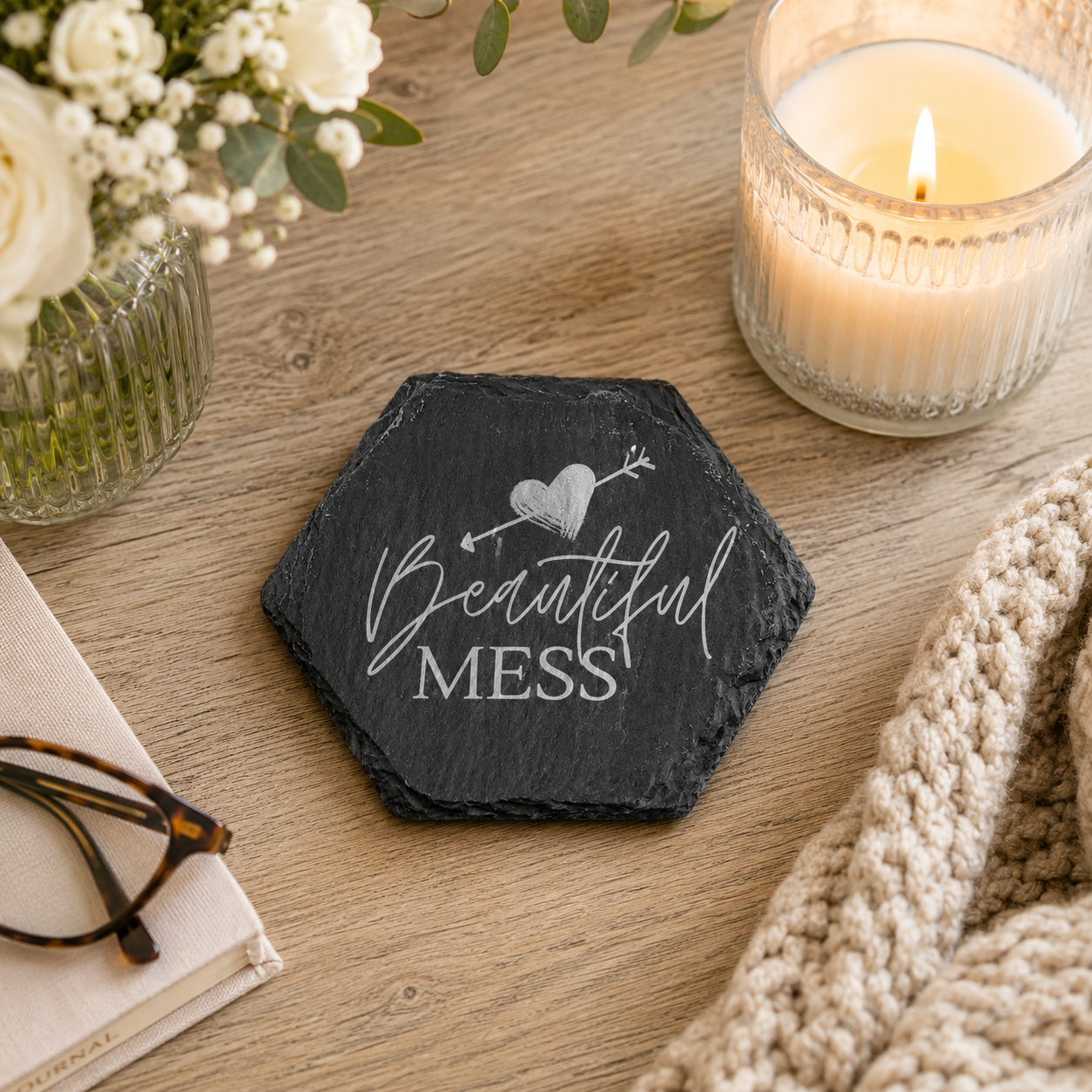 Hexagonal slate coaster with 'beautiful mess' text on a wooden surface with plants, candle and books.