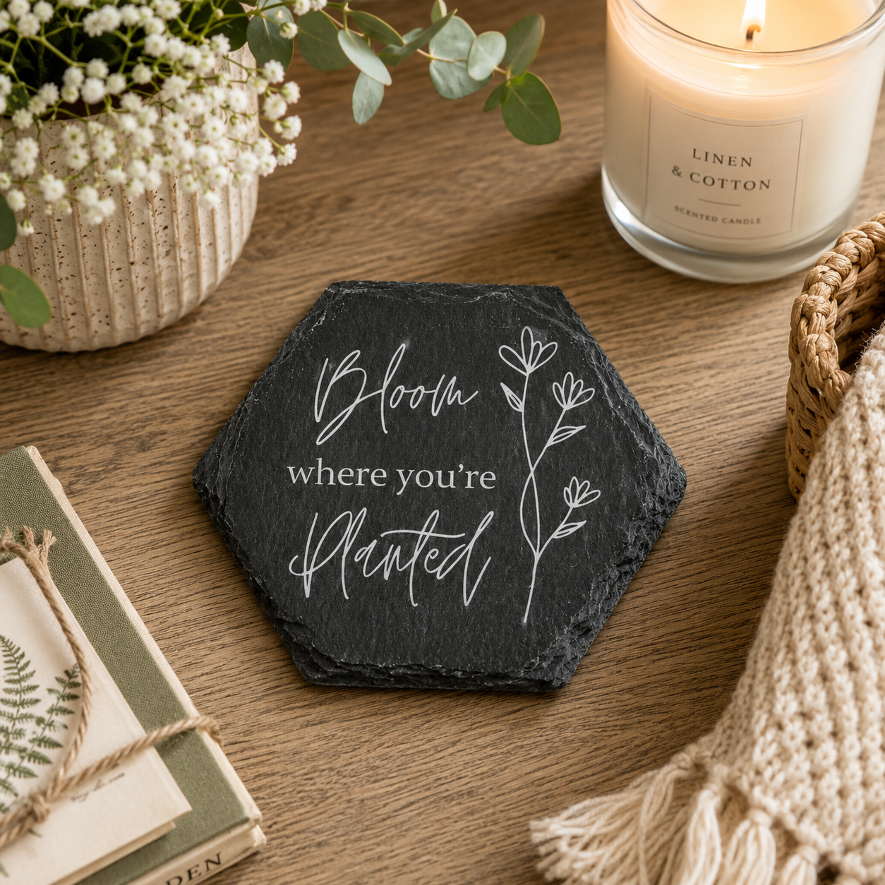 Hexagonal slate coaster with 'Bloom where you're planted' text on a wooden surface with plants and a candle.