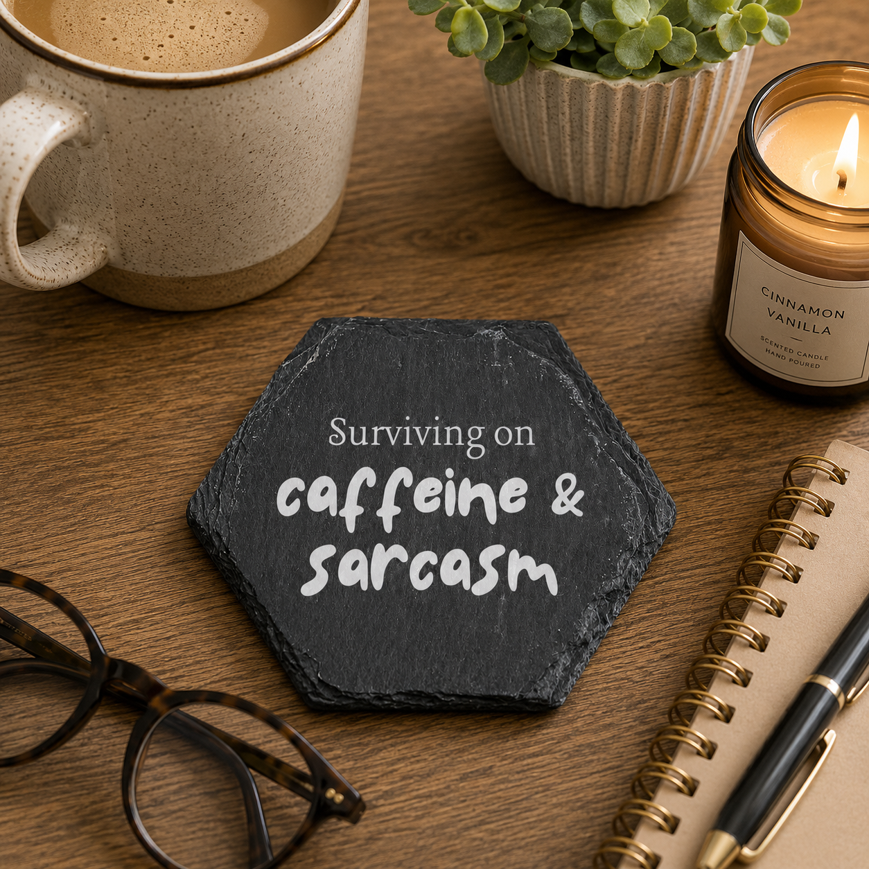 Hexagonal slate coaster with 'Survivng on caffeine and sarcasm' text on a wooden surface with plants, coffee and notebook.