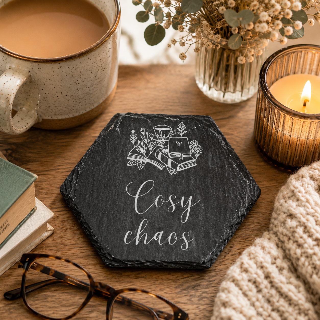 Hexagonal slate coaster with 'cosy chaos' text on a wooden surface with plants, tea, books and glasses.