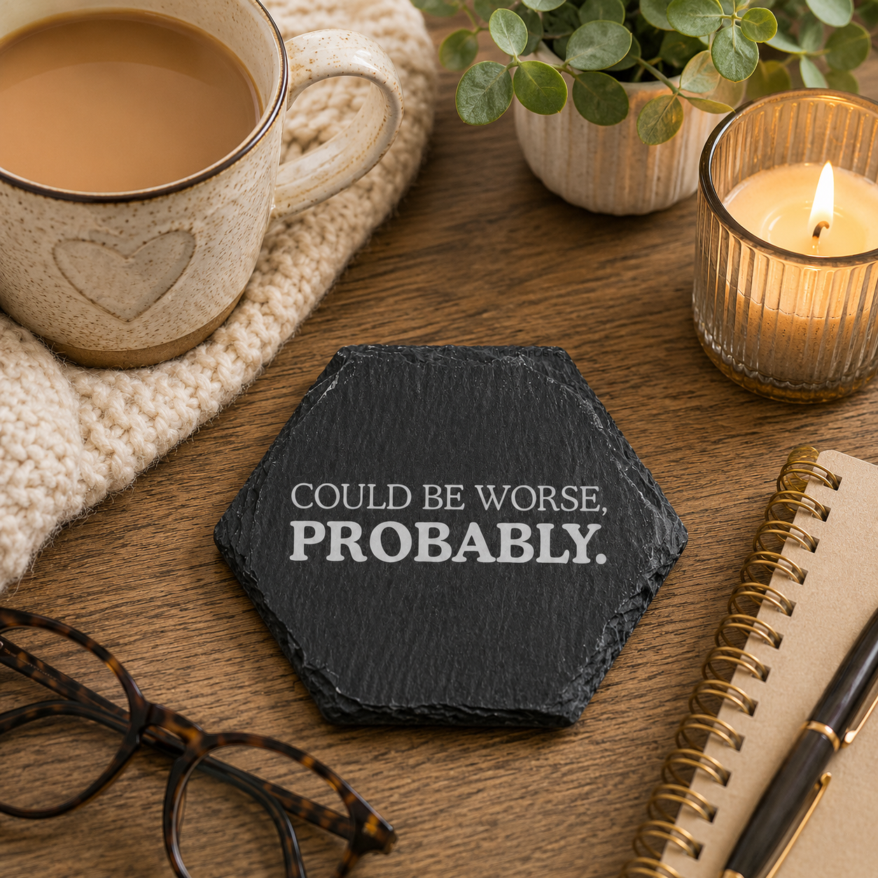 Hexagonal slate coaster with 'could be worse, probably.' text on a wooden surface with plants, tea, glasses and notebook.