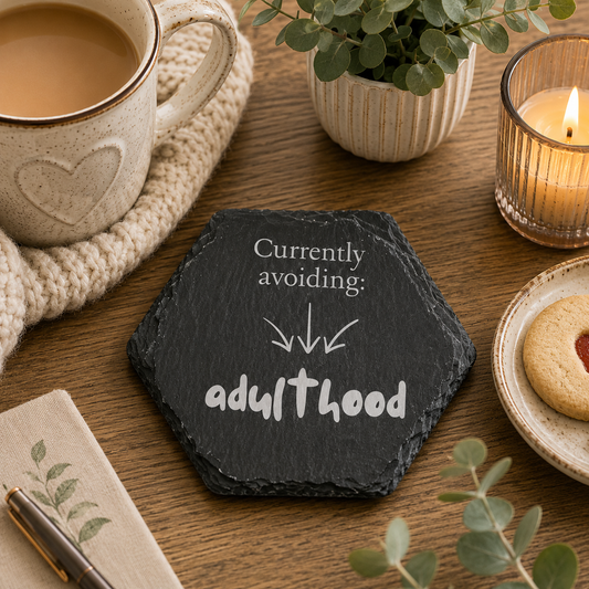 Hexagonal slate coaster with 'currently avoiding: adulthood' text on a wooden surface with plants, tea and biscuits.