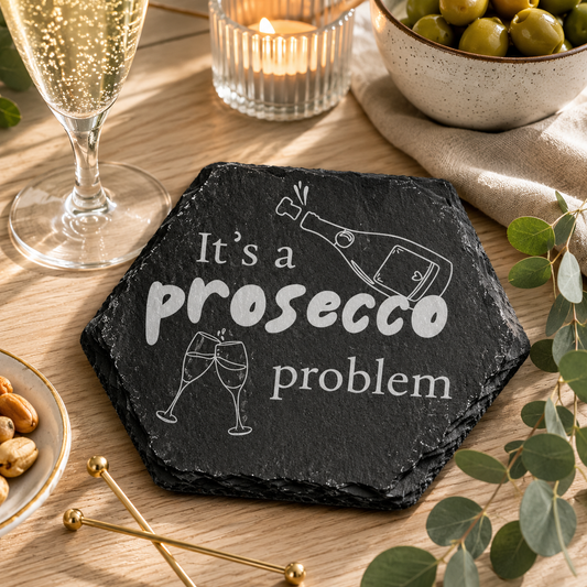 Hexagonal slate coaster with 'it's a prosecco problem' text on a wooden surface with a prosecco glass, olives and nuts.