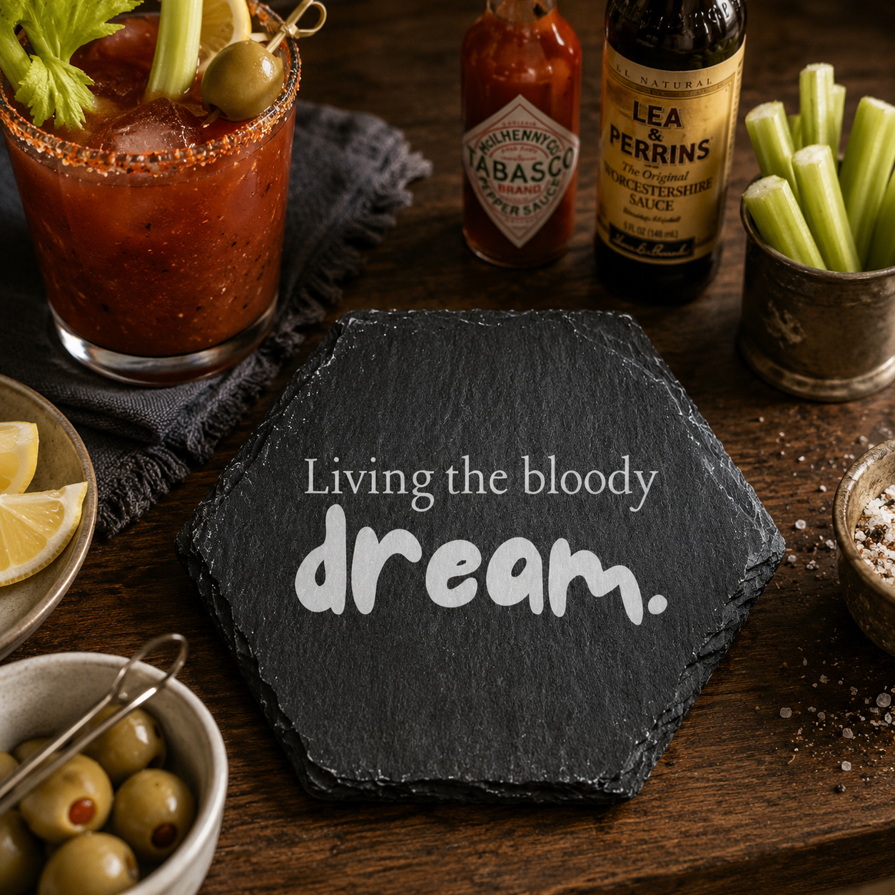 Hexagonal slate coaster with 'living the bloody dream' text on a wooden surface with cocktail glass, bottles and accesories.
