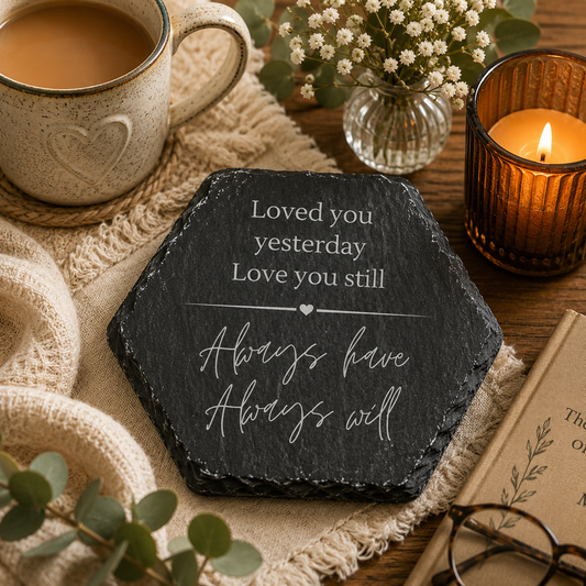 Hexagonal slate coaster with a romantic quote on a wooden surface with plants, books and candles.