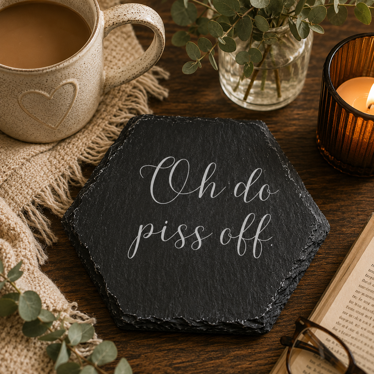 Hexagonal slate coaster with 'oh do piss off' text on a wooden surface with plants, tea and candles.