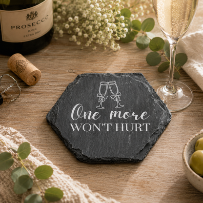 Hexagonal slate coaster with 'one more won't hurt' text on a wooden bar with prosecco bottle and glasses.