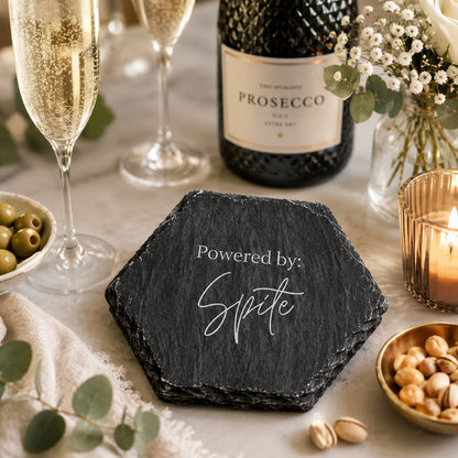 Hexagonal slate coaster with 'Powered by: Spite' text on a marble surface with prosecco bottle and glasses.