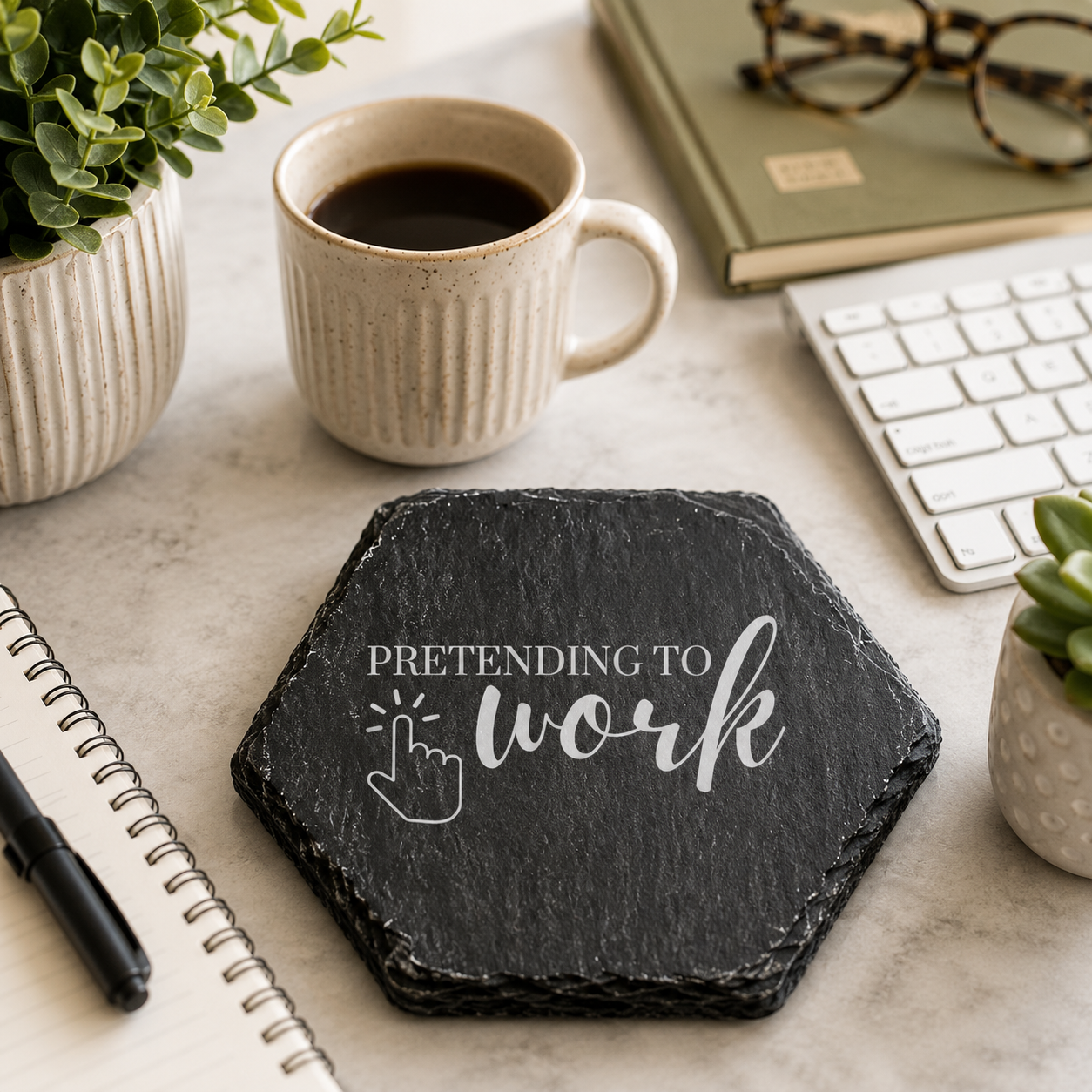 Hexagonal slate coaster with 'pretending to work' text on a marble desk with keyboard, notebook and coffee.