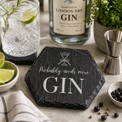 Hexagonal slate coaster with 'probably needs more gin' text on a marble surface with gin bottle and glasses.