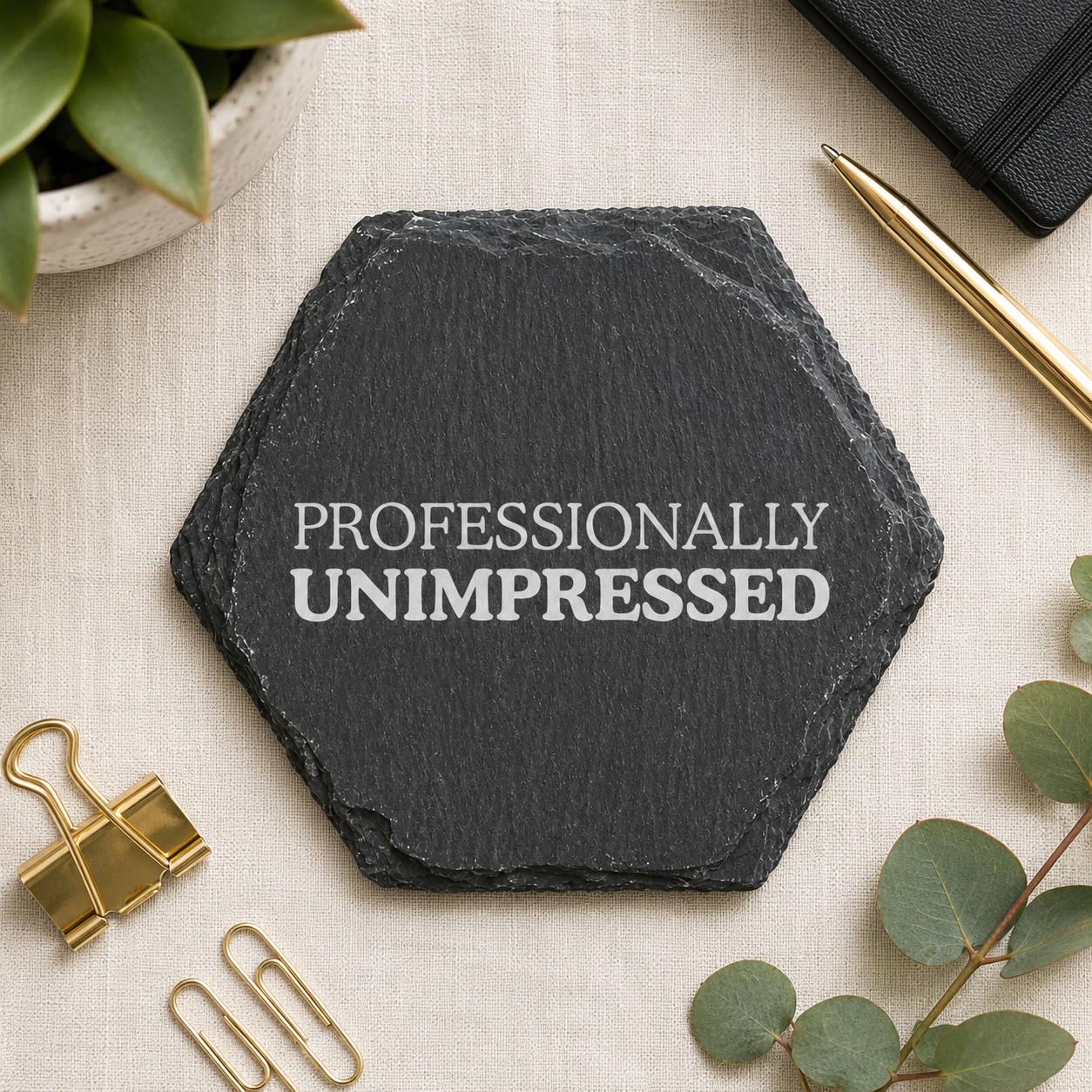 Hexagonal black slate coaster with 'PROFESSIONALLY UNIMPRESSED' text on a light surface with office supplies.