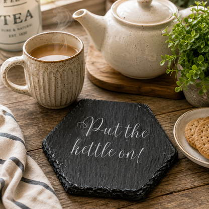 Hexagonal slate coaster with 'put the kettle on' text on a wooden surface with plants, teapot and teacup.