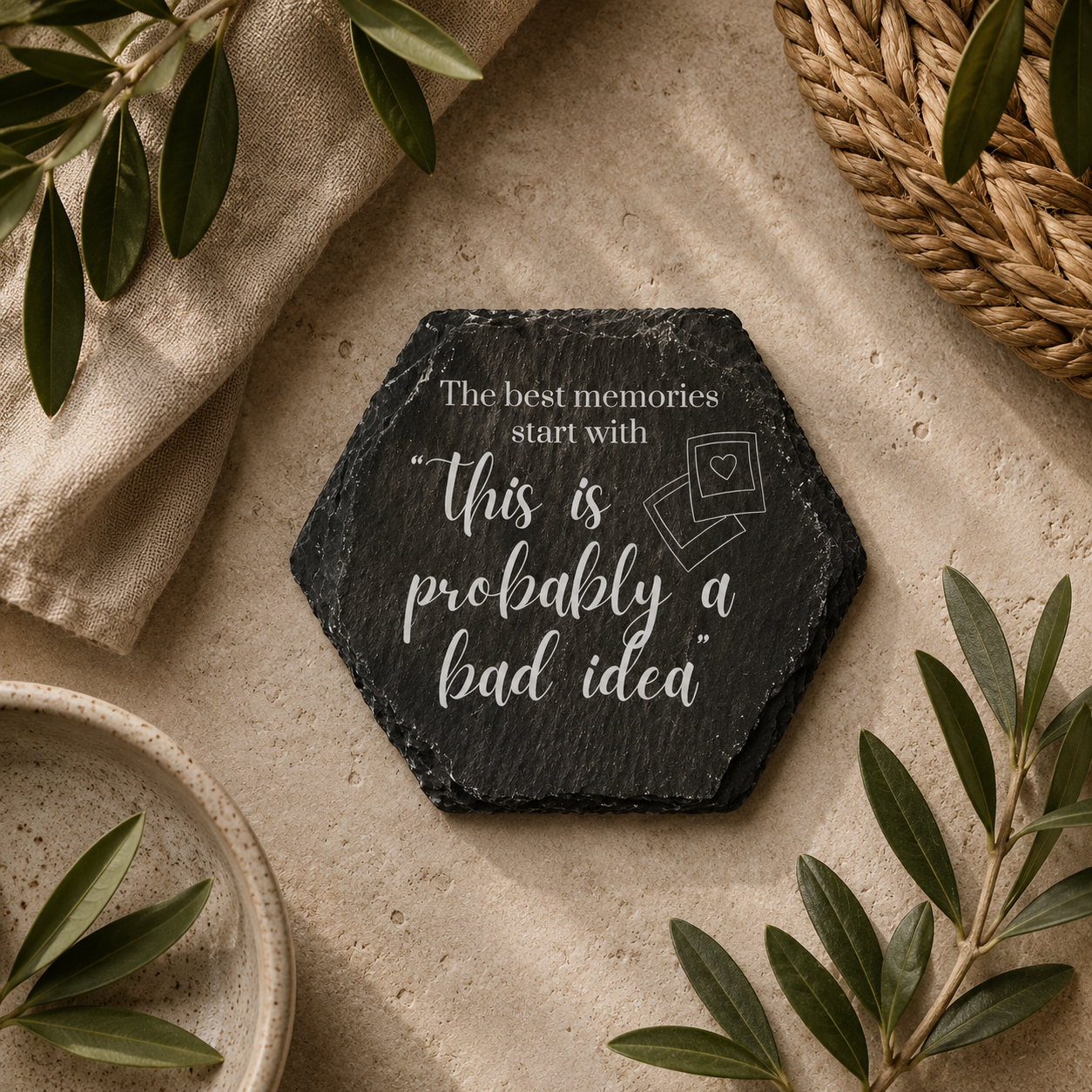 Hexagonal slate coaster with 'the best memories start with this is probably a bad idea' text on a wooden surface with plants.