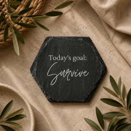 Hexagonal slate coaster with 'today's goal: survive' text on a wooden surface with plants.