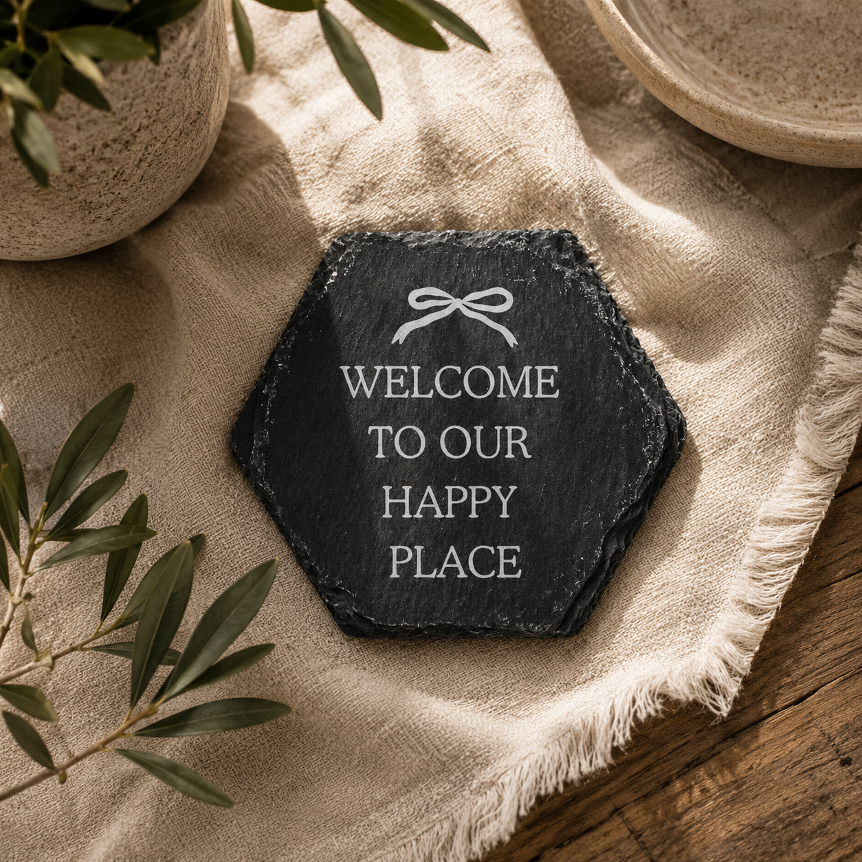 Hexagonal slate coaster with 'welcome to our happy place' text on a wooden surface with plants.