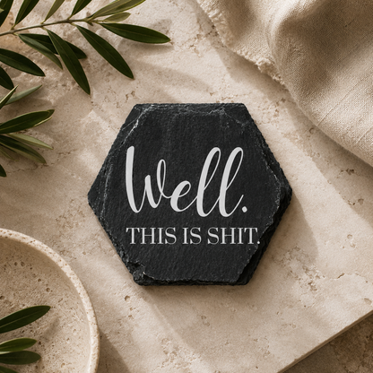 Hexagonal black slate coaster with white 'well this is shit' text on a beige surface with green leaves.