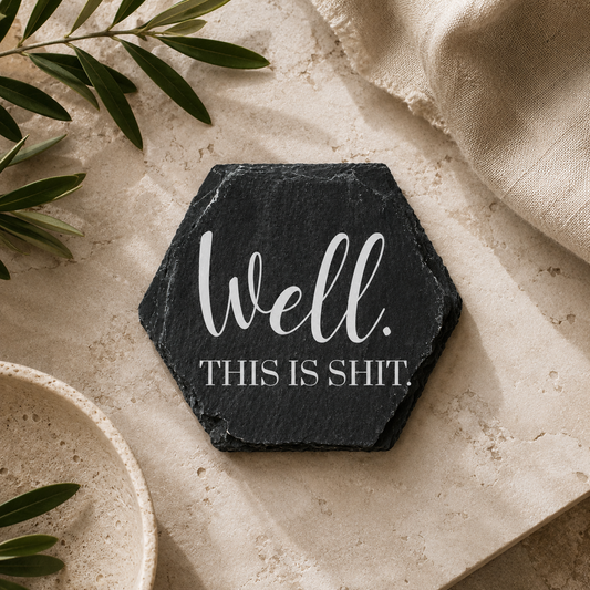 Hexagonal black slate coaster with white 'well this is shit' text on a beige surface with green leaves.