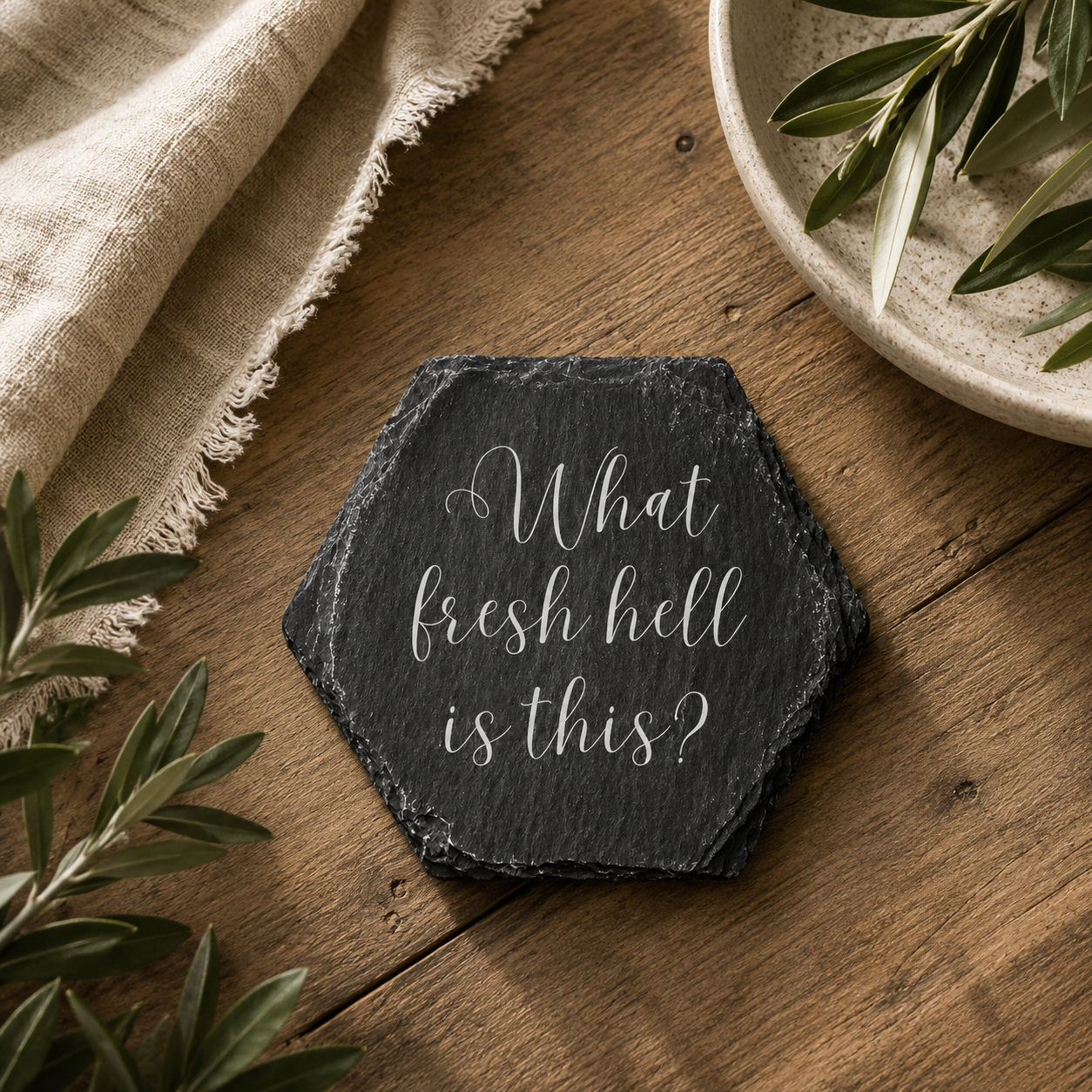 Hexagonal slate coaster with 'what fresh hell is this' text on a wooden surface with plants.