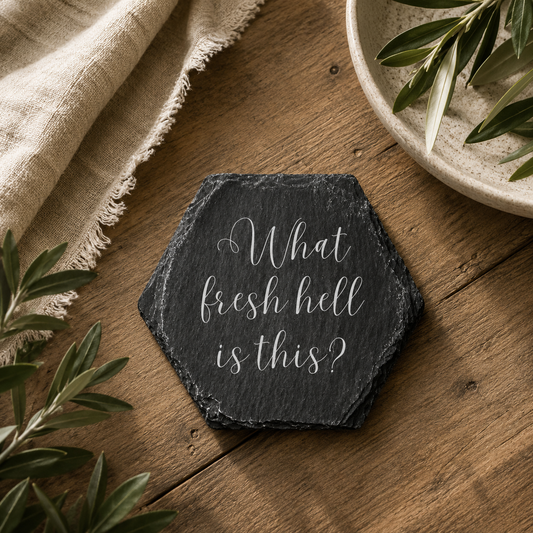 Hexagonal slate coaster with 'what fresh hell is this' text on a wooden surface with plants.