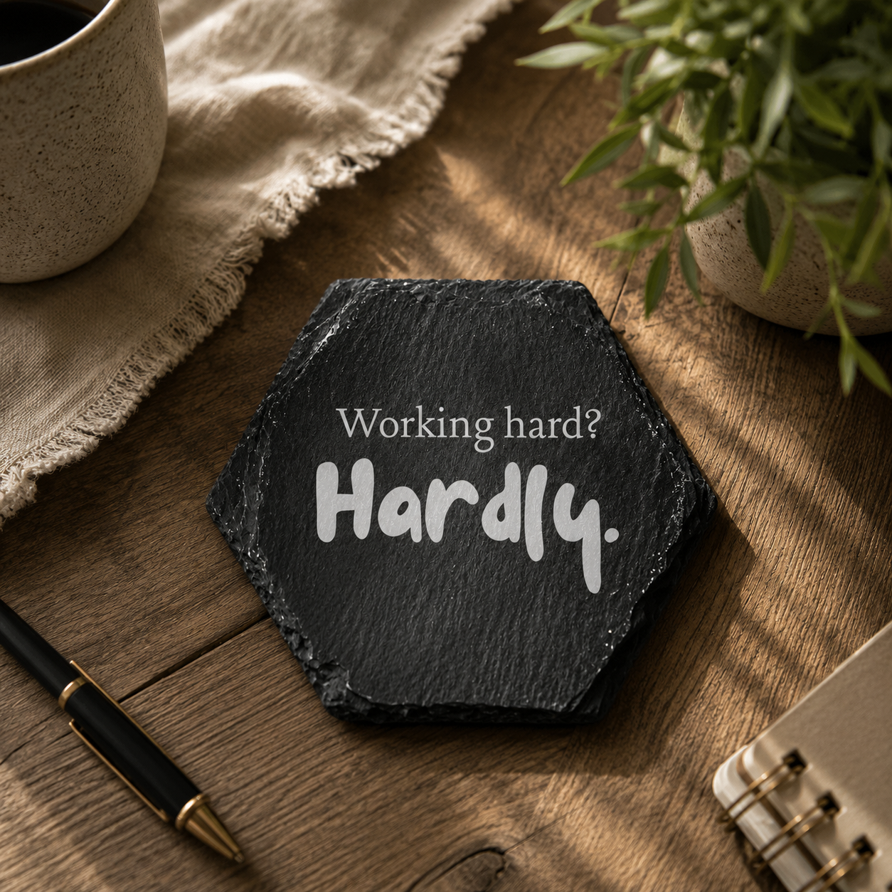 Hexagonal slate coaster with 'working hard? hardly.' text on a wooden surface with plants.