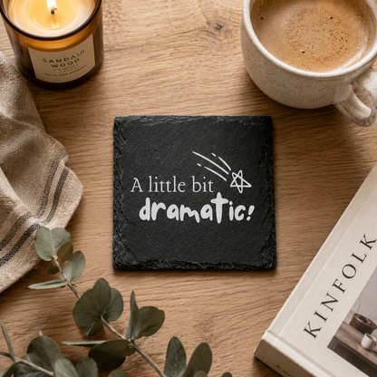 Slate coaster with 'A little bit dramatic!' text on a wooden table with a candle, mug, and book.