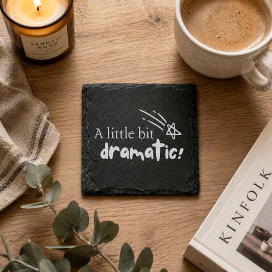 Slate coaster with 'A little bit dramatic!' text on a wooden table with a candle, mug, and book.