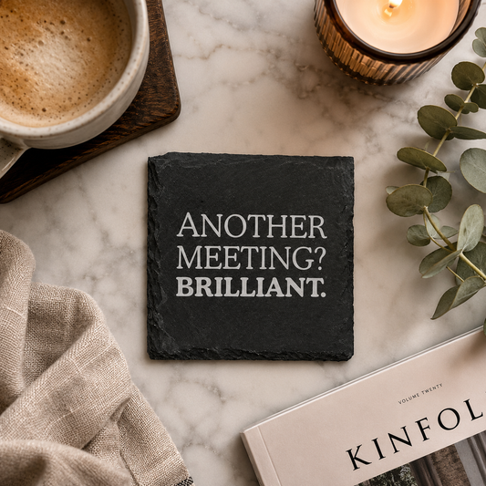 Slate coaster with 'Another meeting? Brilliant.' text on a marble surface with coffee, book, and candle.