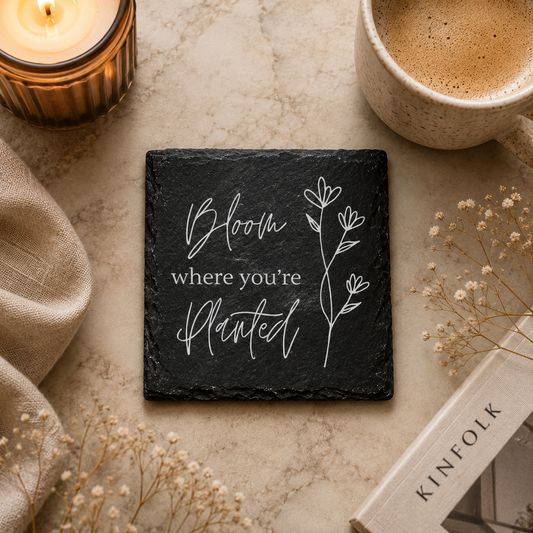 Slate coaster with 'Bloom where you're planted' quote on a table with coffee, candle, and book.
