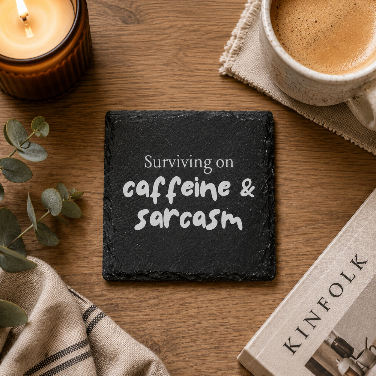 Slate coaster with 'Surviving on caffeine and sarcasm' text on a wooden table with a cup of coffee, candle, and book.