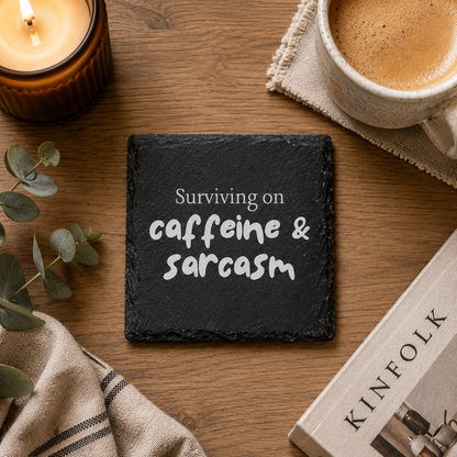 Slate coaster with 'Surviving on caffeine and sarcasm' text on a wooden table with a cup of coffee, candle, and book.