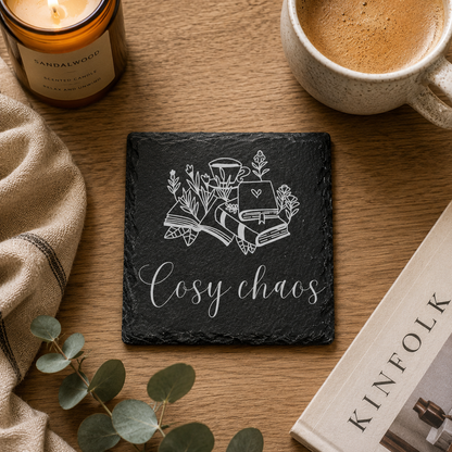 Slate coaster with 'Cosy chaos' text and illustrations on a wooden surface with a candle, mug, and book.