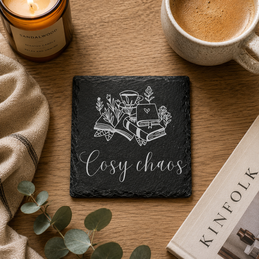Slate coaster with 'Cosy chaos' text and illustrations on a wooden surface with a candle, mug, and book.