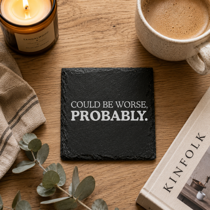 Slate coaster with 'Could be worse, probably' text on a wooden table with coffee, candle, and book.
