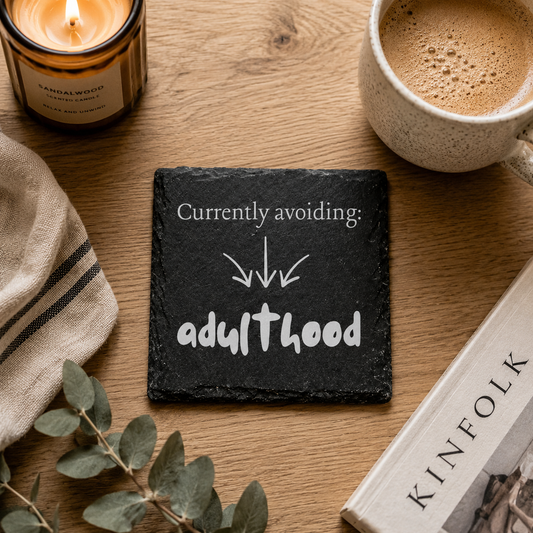 Slate coaster with 'Currently avoiding: adulthood' text on a wooden table with a cup of coffee, candle, and book.