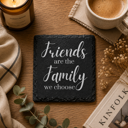 Slate coaster with 'Friends are the family we choose' quote on a wooden surface with a cup, candle, and book.