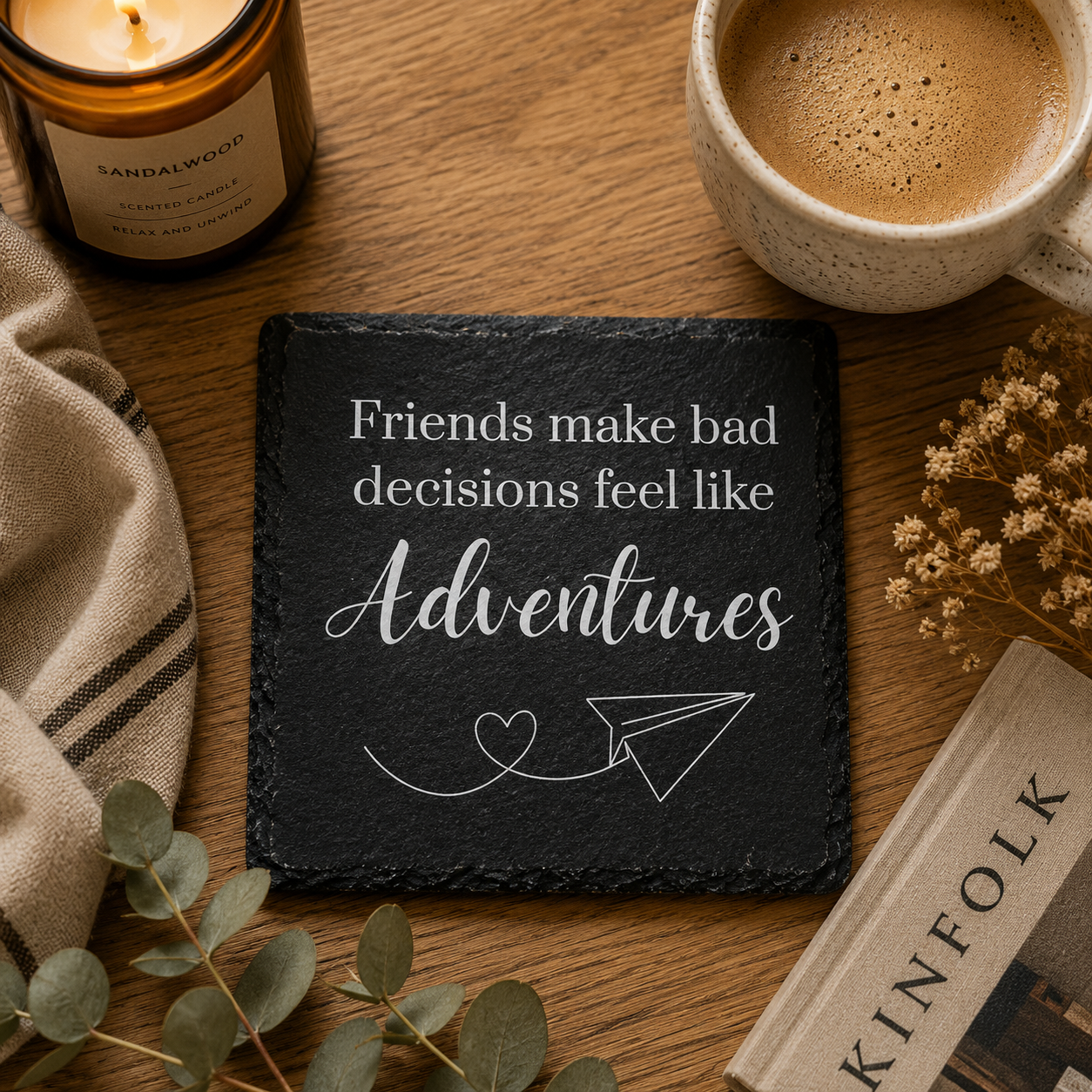 Slate coaster with motivational quote, mug of coffee, candle, and book on wooden surface