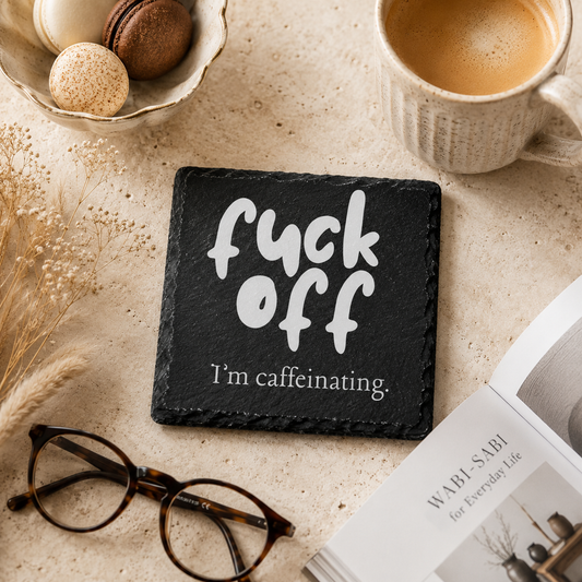 Slate coaster with 'fuck off, I'm caffeinating' text on a table with coffee and glasses.