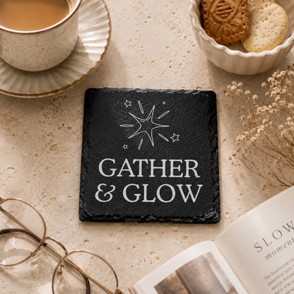 Slate coaster with 'Gather & Glow' text on a table with a cup of coffee, cookies, and glasses.