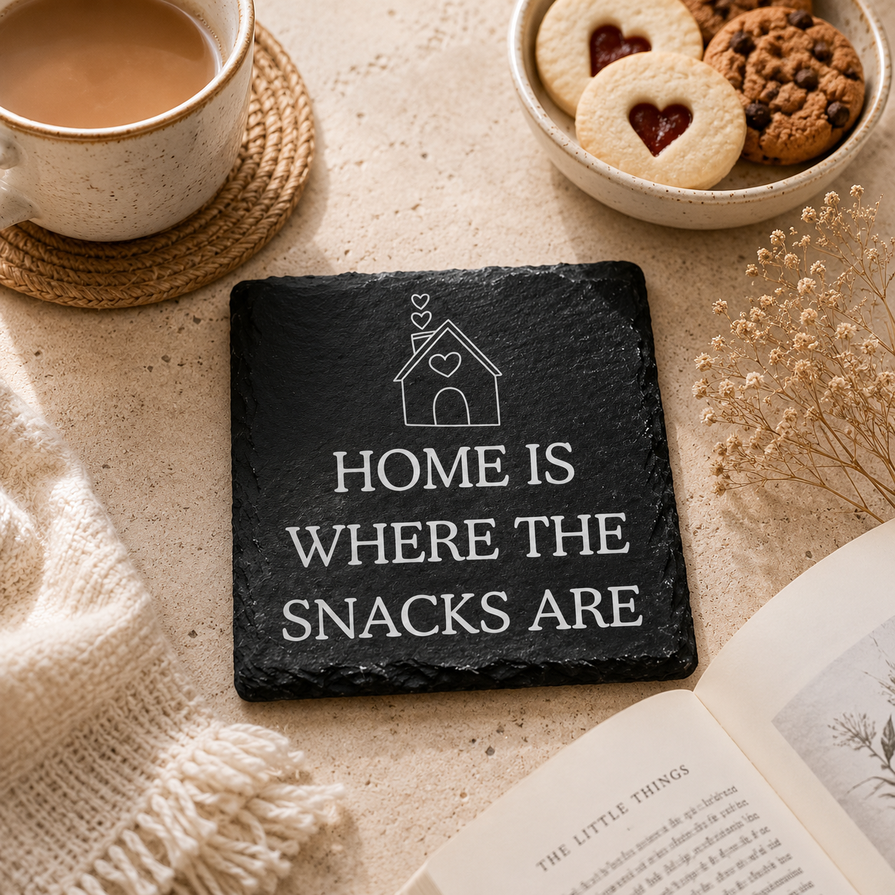 Slate coaster with 'Home is where the snacks are' text on a wooden table with a cup of coffee, candle, and book.