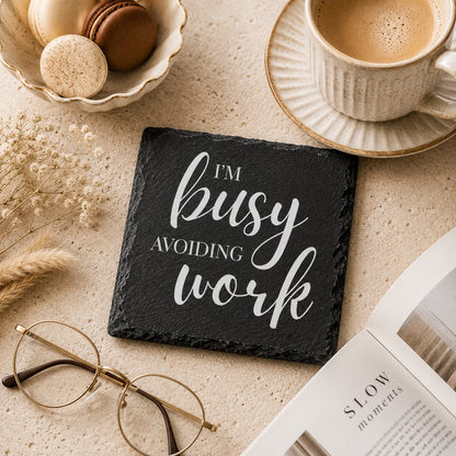 Slate coaster with 'I'm busy avoiding work' text on a table with coffee, glasses, and a book.