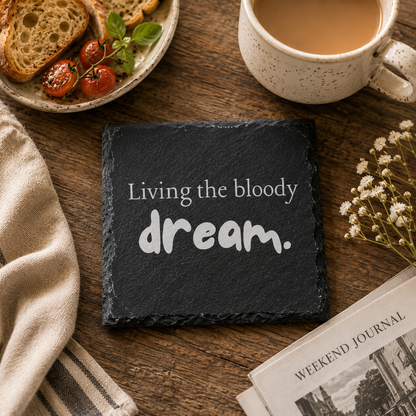 Slate coaster with 'Living the bloody dream' text on a wooden table with coffee, toast, and flowers.