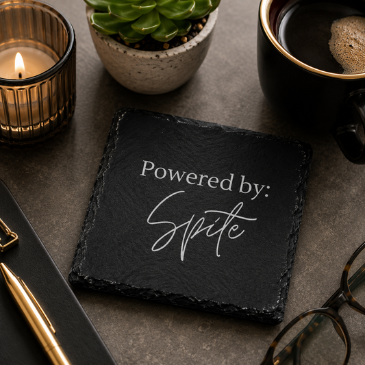 Black slate coaster with 'Powered by: Spite' text on a table with a candle, plant, and mug.