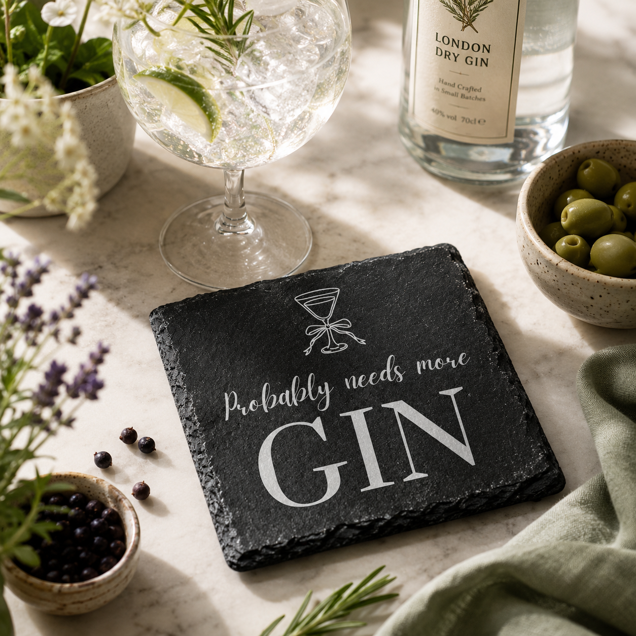 Slate coaster with 'Probably needs more Gin' text on a table with gin and tonic setup.