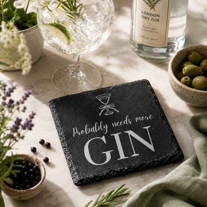Slate coaster with 'Probably needs more Gin' text on a table with gin and tonic setup.