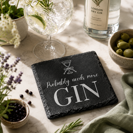 Slate coaster with 'Probably needs more Gin' text on a table with gin and tonic setup.