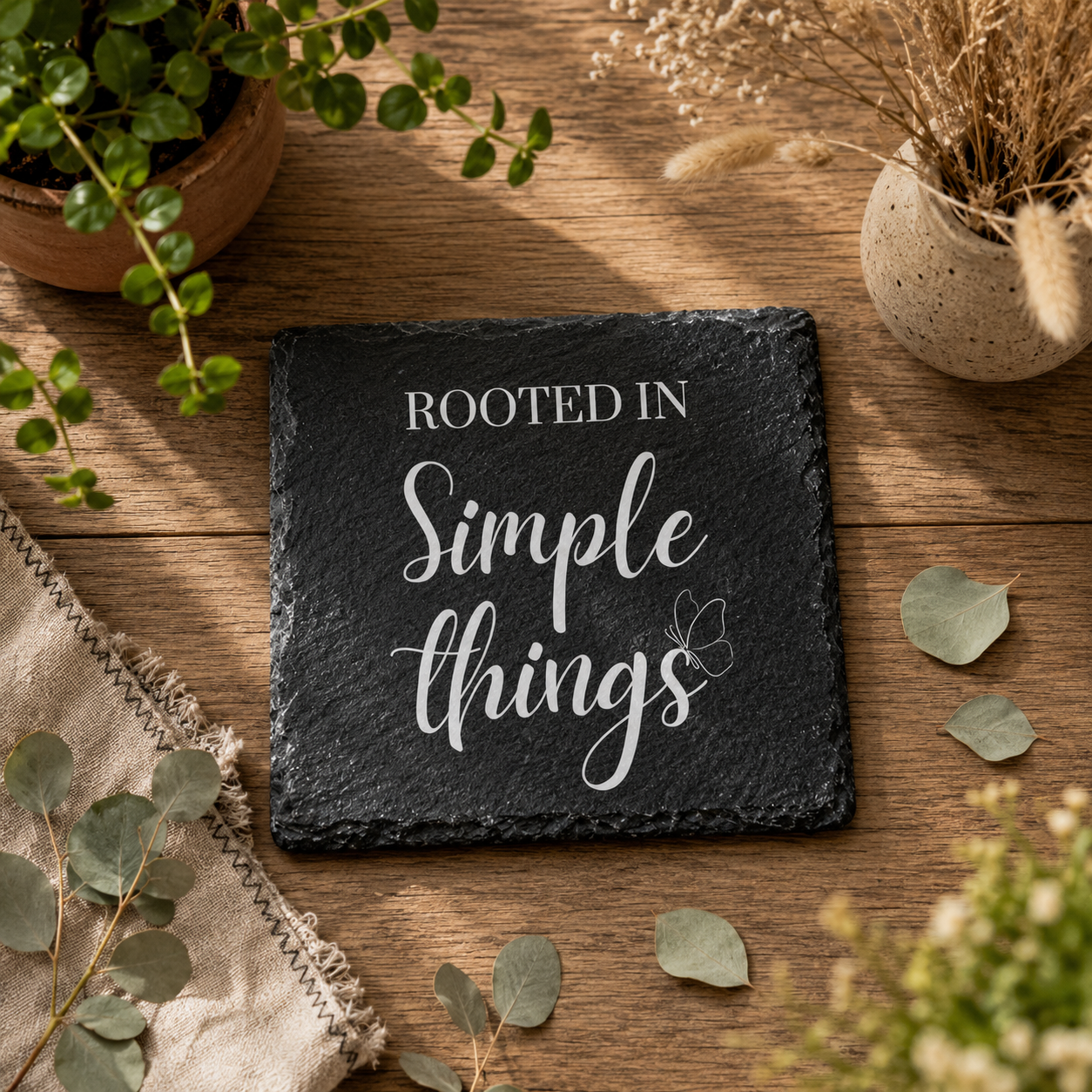 Slate coaster with 'Rooted in Simple Things' text on a wooden surface with plants.