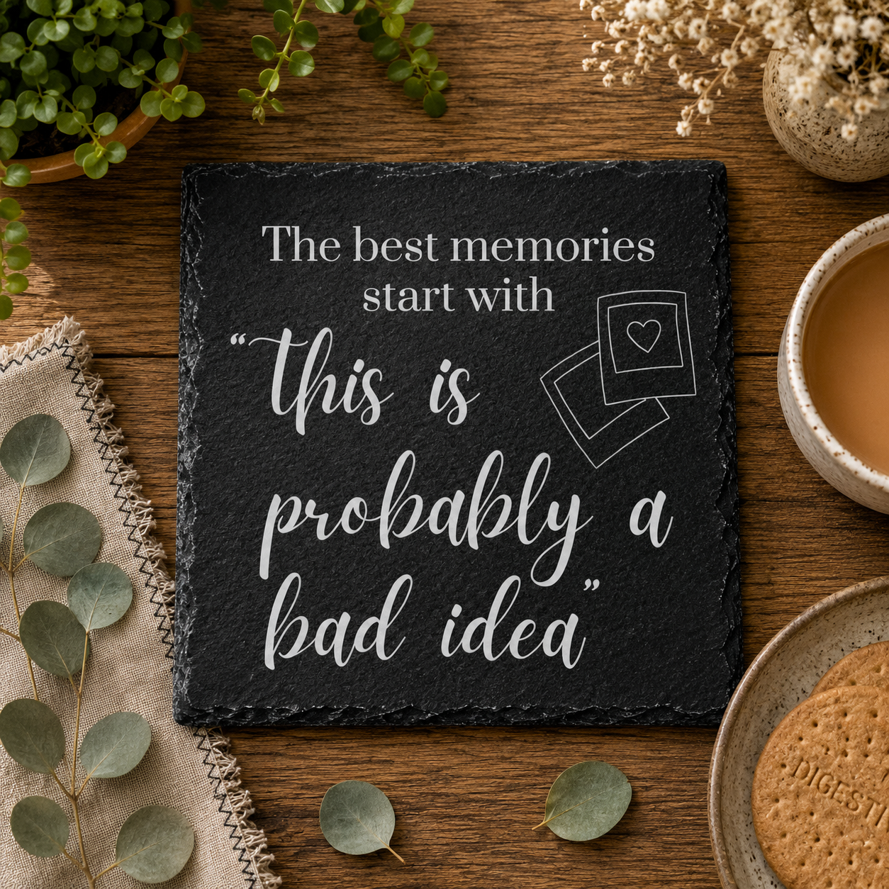 Slate coaster with humorous quote on a wooden table with plants and a cup of tea.