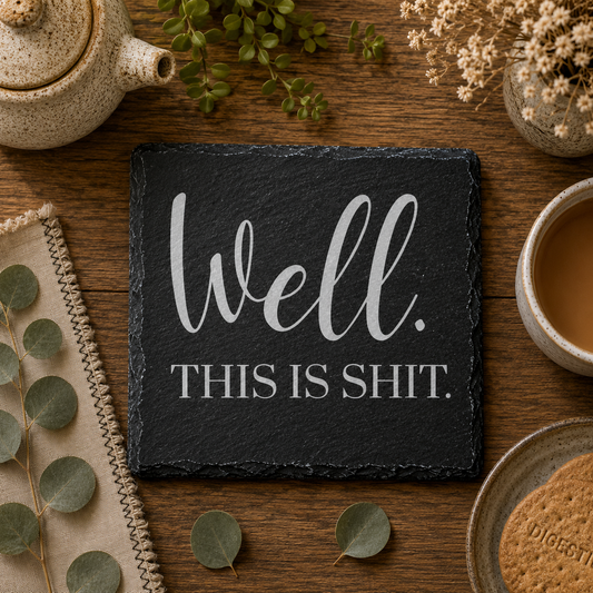 Slate coaster with 'Well. This is shit.' text on a wooden table with tea and plants.