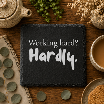 Slate coaster with 'Working hard? Hardly.' text on a wooden table with tea and plants.